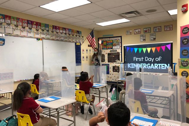 students sit at a desks with three-sided barriers and wear masks as their teacher wears a mask at the front of the classroom